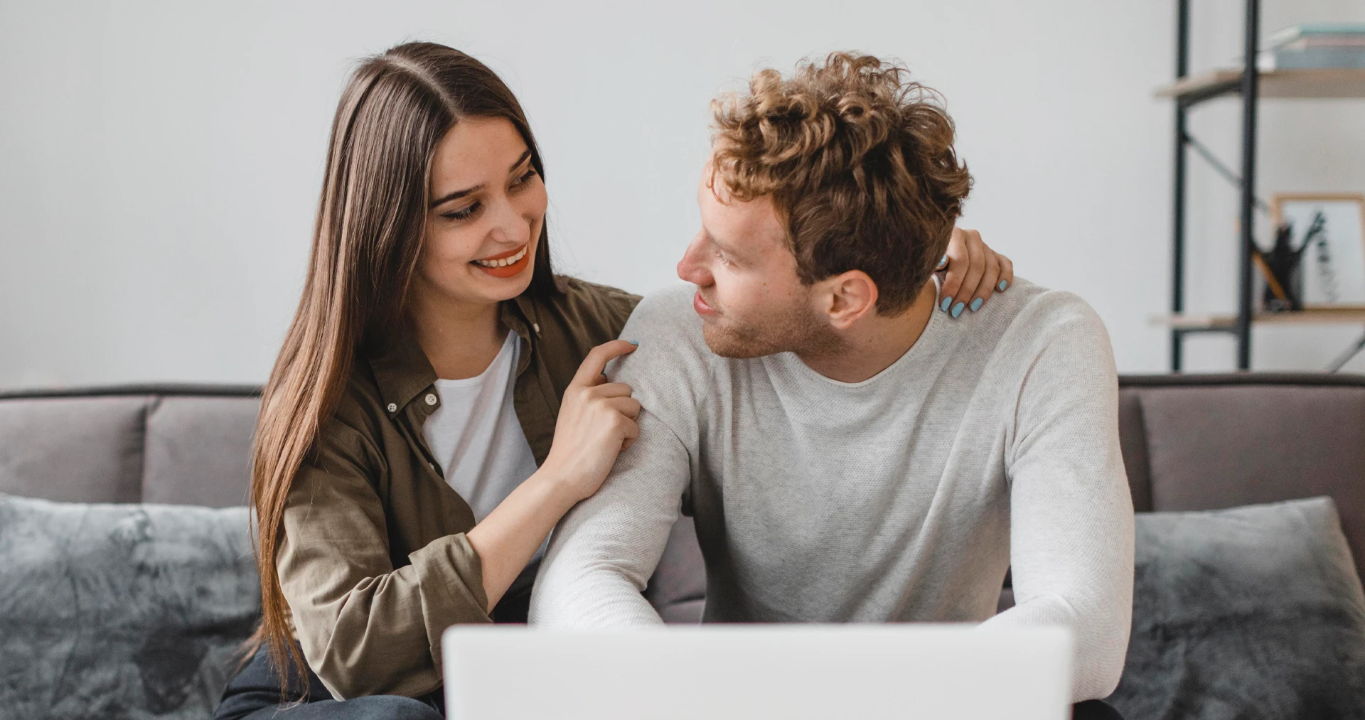 Fiatal pár ül a kanapén laptop előtt, egymásra mosolyogva beszélik át pénzügyeiket
