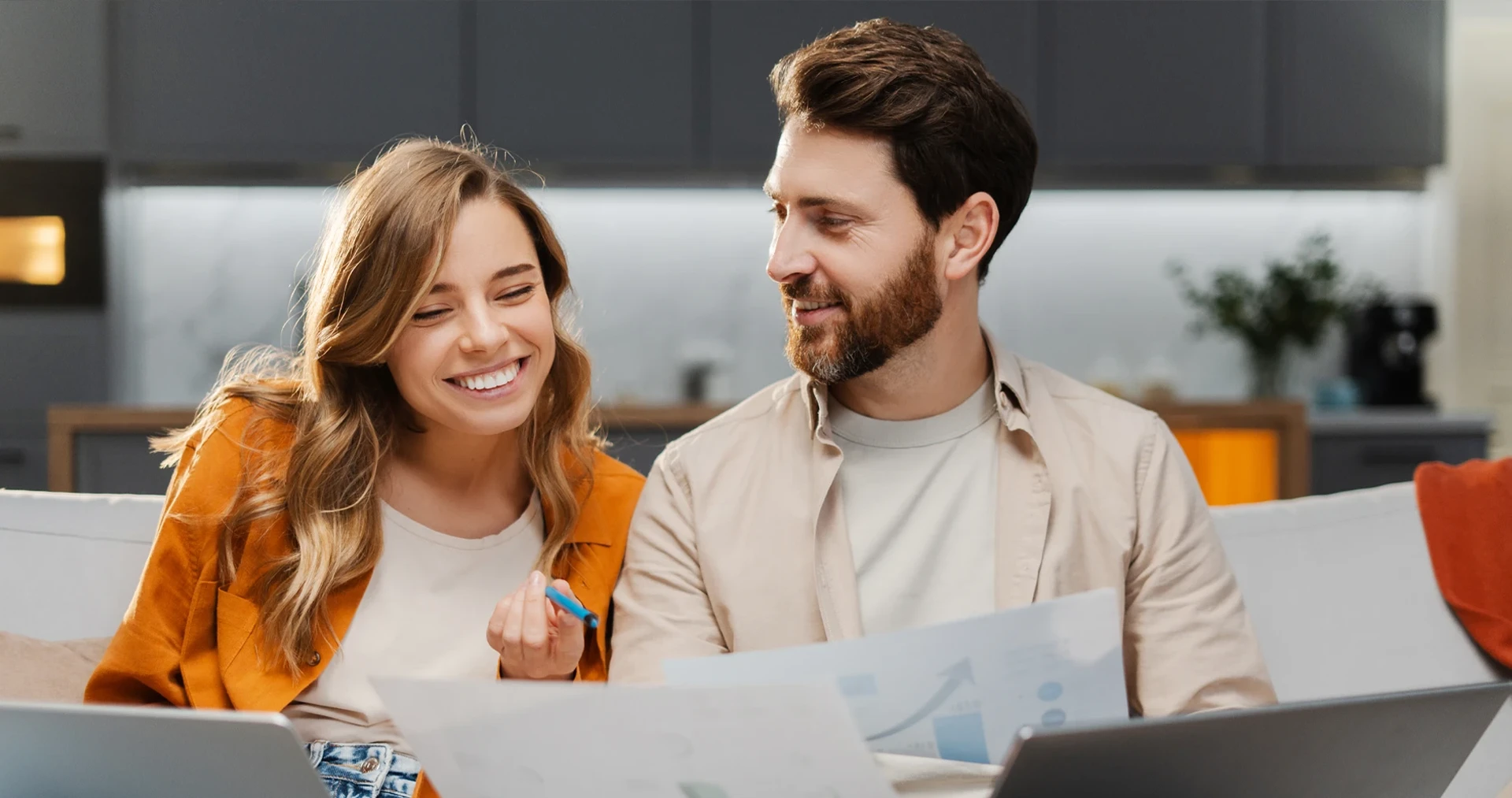 Mosolygó pár ül az asztalnál, laptop és papírok fölé hajolva, közösen áttekintik a hitelügyintézés dokumentumait szakértői támogatással.